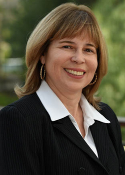 Woman with brown hair wearing a black coat over a white blouse and hoop earrings