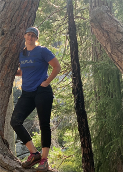 Woman wearing a blue t-shirt, black pants and a grey cap standing next to a tree